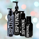 Four bottles of SPUNK Hybrid Lubricant from Spunk Lube, known for their hypoallergenic formula, are displayed against a blurred backdrop with multicolored lights. These black bottles with white text are available in different sizes and feature various pump and cap styles, providing the ultimate water-based silicone lubrication experience.